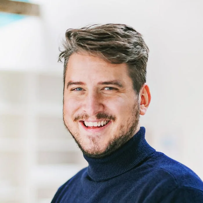 Philipp Maxhofer — Le Werk, Köln Philipp Maxhofer, Creative Director bei Le Werk in Köln, im dunkelblauen Rollkragenpullover, lachend vor den hellen Regalen des Studios