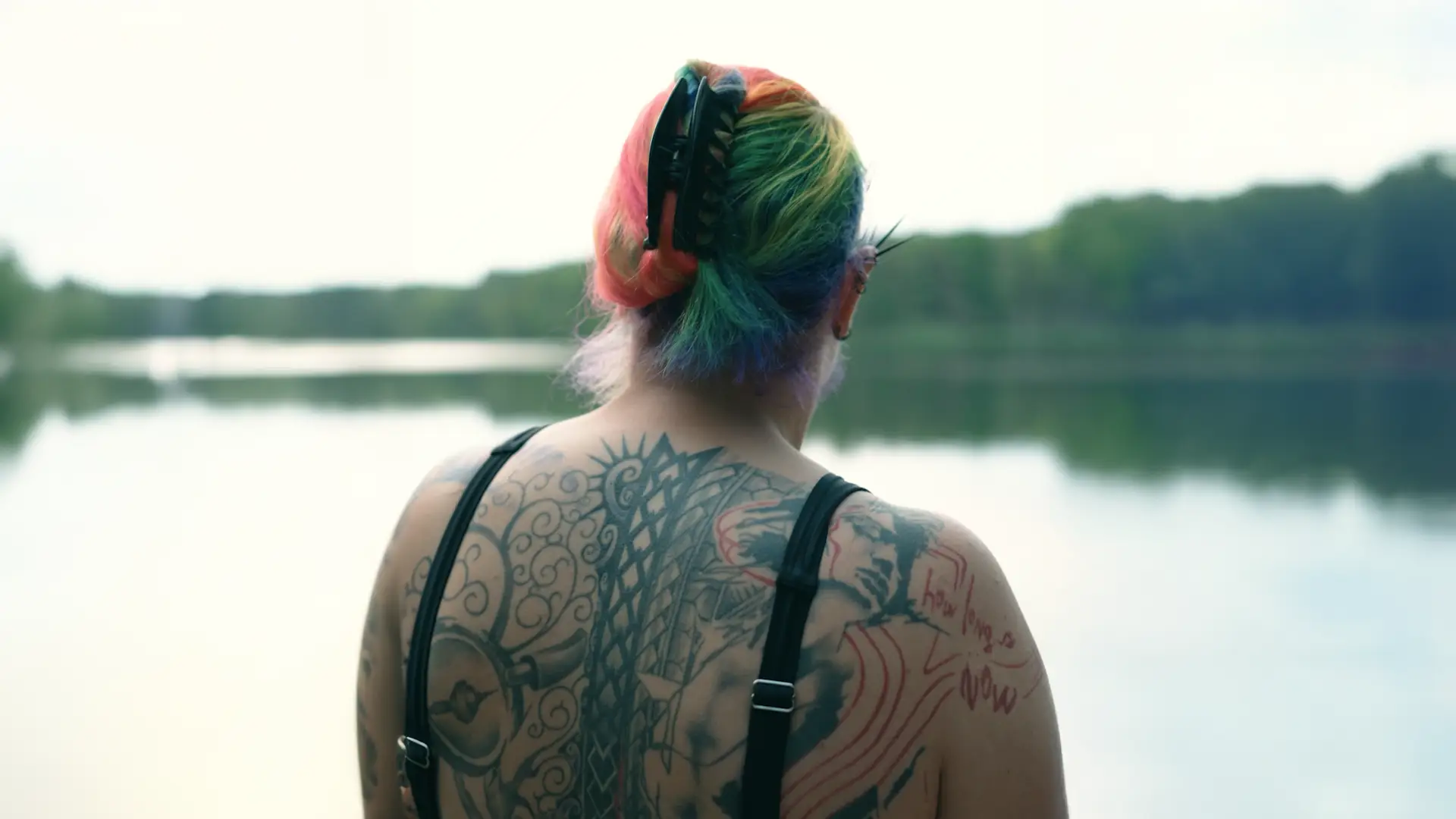 Mitarbeiterporträt für Siemens Standbild aus einem Mitarbeiterporträt für Siemens: Frau mit regenbogenbunt gefärbtem Haar und großflächigem Rückentattoo, von hinten am Ufer eines Sees