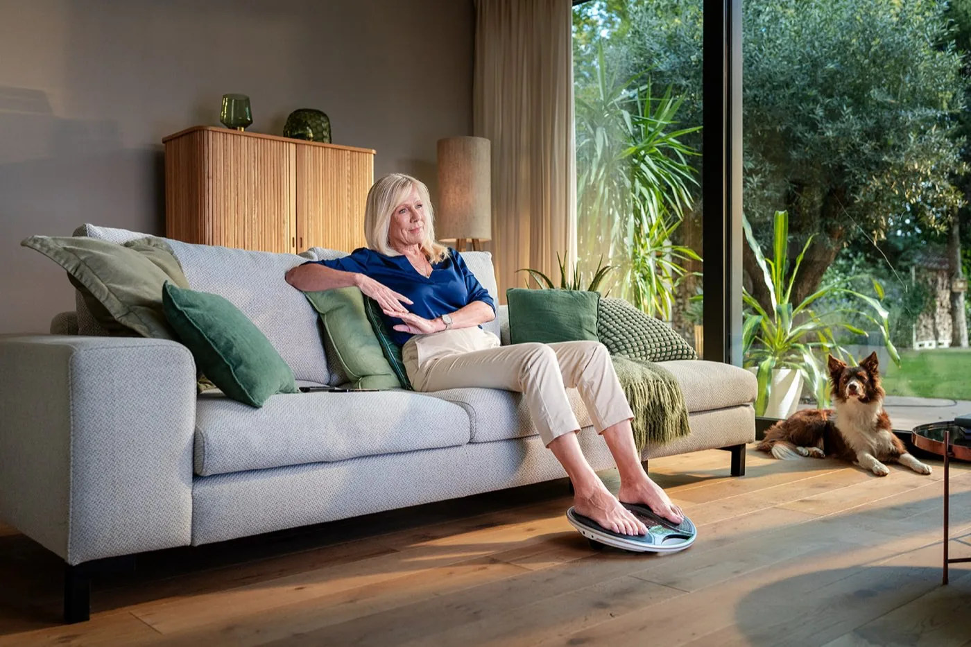 Aus der Produktfotografie für Revitive: Ulrike von der Groeben sitzt auf dem hellen Sofa, die Füße auf dem Durchblutungsgerät, daneben liegt der Hund